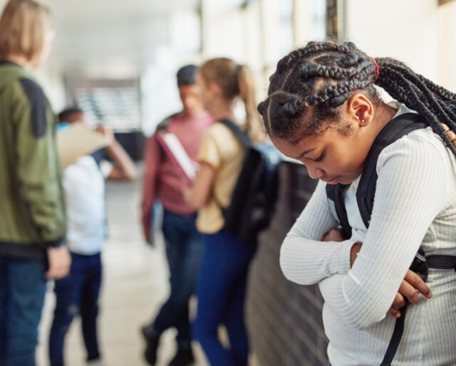 Shot of a young girl looking sad while being excluded from her peers in the hallway of a school