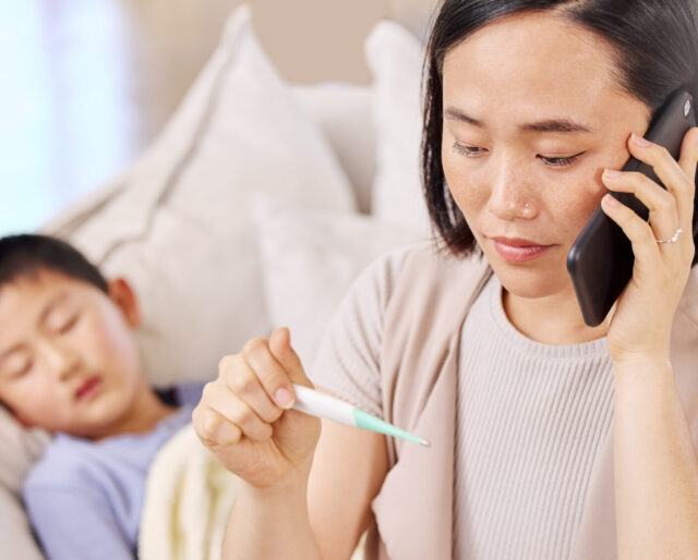 mother on phone looking at a thermometer while her sleeping child lays in bed behind her