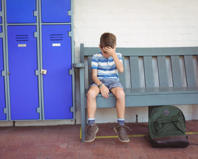 Full length of sad boy sitting on bench by lockers at school