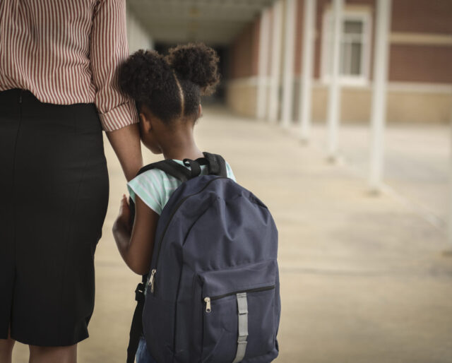 Young girl with backpack leaning on arm of adult at school.